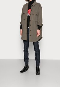 Donna che indossa una giacca verde oliva con bottoni, un maglione nero con design di rosa rossa, jeans scuri e stivaletti neri contro uno sfondo bianco.