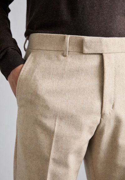 Beige uldbukser med en glat tekstur, der har en skræddersyet pasform, sidelommer og en flad front. Brun sweater synlig ovenfor.