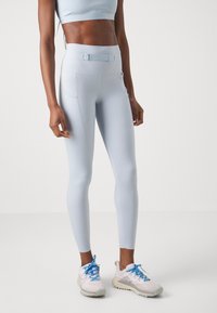 Világoskék, magas derekú leggings sima textúrával, oldal zsebekkel és elülső záróhevederrel, világos sportcipőkkel párosítva.
