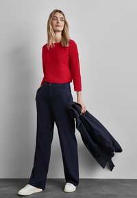 Top rosso a maniche lunghe con finitura testurizzata, abbinato a pantaloni larghi blu navy. Il modello tiene in mano un cappotto blu navy; le sneakers bianche completano l'outfit.