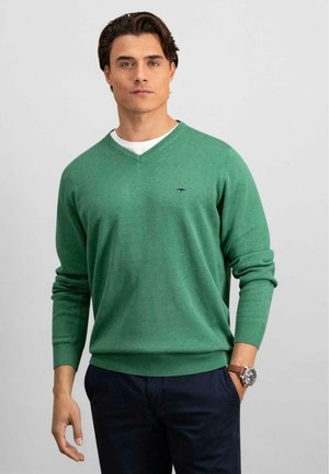 Green v-neck sweater with long sleeves, ribbed cuffs and hem, worn over a white t-shirt, paired with dark trousers. Subtle logo detail on chest.