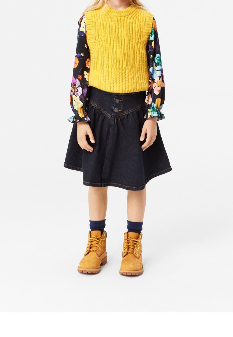 Enfant portant un gilet jaune en tricot, une chemise à manches longues fleurie, une jupe en jean foncé, des chaussettes bleu marine et des bottines lacées beige, debout sur fond blanc.