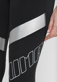 Černé sportovní legíny mají texturovaný povrch, široký stříbrný pruh na boku a výrazný šedý logo design na spodní části nohy.