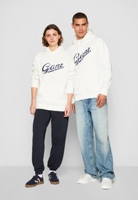Hvite hettegensere med en marineblå "Gant"-logo, kombinert med marineblå joggebukser og lyseblå jeans. Avslappet, komfortabel design med snorer.
