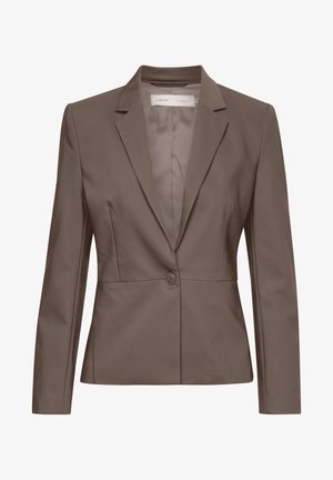 Vrouw met een taupekleurig pak met cropped broek en zwarte slip-on schoenen, zijwaarts staand tegen een effen witte achtergrond.