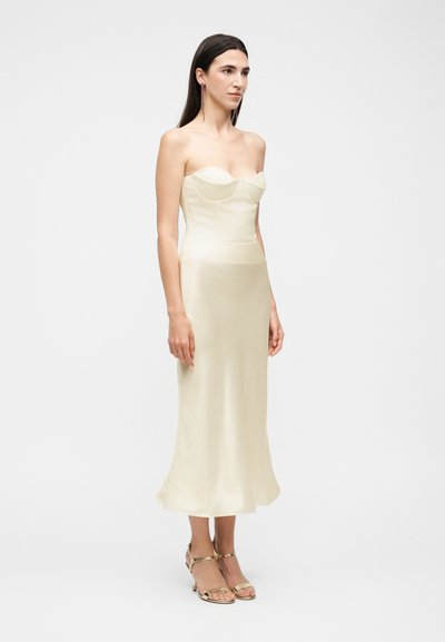 Femme aux longs cheveux foncés portant une robe midi en satin crème bustier et des talons ouverts dorés, debout devant un fond blanc uni.