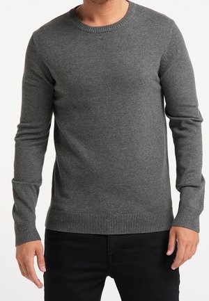 Mann trägt einen eng anliegenden, langärmeligen, dunkelgrauen Strickpullover mit gerippten Bündchen und Saum, kombiniert mit schwarzen Hosen.