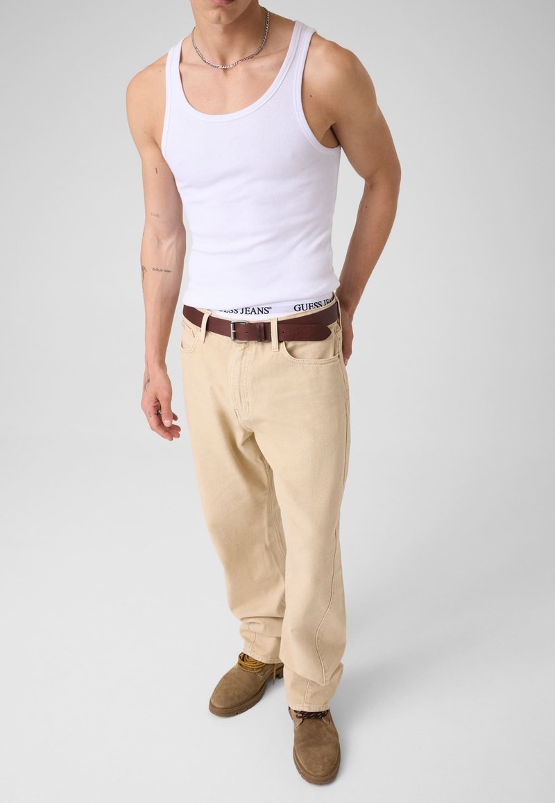 Homme portant un débardeur blanc, un pantalon beige, une ceinture marron, des chaussures marron et une ceinture blanche visible avec le texte "GUESS JEANS", debout devant un fond uni.