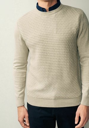 Mand iført beige tekstureret striktrøje over mørkeblå skjorte med krave, med mørke bukser, vist fra nakke til hofter.