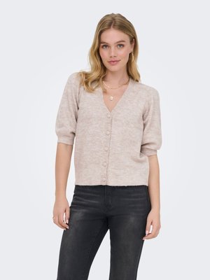 Femme portant un cardigan beige boutonné avec des manches courtes bouffantes et un jean noir, debout devant un fond clair uni.