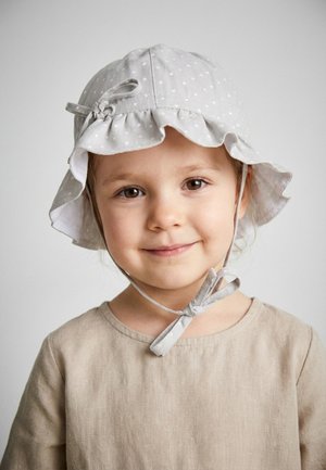Lächendes kleines Kind mit einem hellgrauen Sonnenhut mit weißen Punkten und Kinnbändern sowie einem beigefarbenen Leinenkleid vor schlichter Hintergrund.