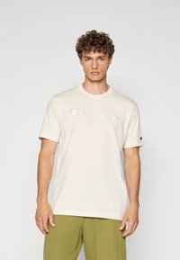 Cremefarbiges Baumwoll-T-Shirt mit rundem Halsausschnitt, kurzen Ärmeln und gesticktem "Champion"-Logo. Kombiniert mit olivgrünen Hosen.