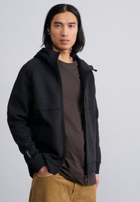 Svart zip-up hettegenser med en jevn tekstur, med foret hette, frontlommer og kontrasterende sømmer. Båret over en mørkebrune skjorte.