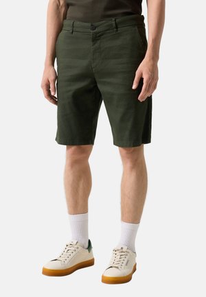 Mężczyzna noszący ciemnozielone krótkie spodenki do kolan, białe sportowe skarpetki i białe trampki z gumowymi podeszwami, stojący na jednolitym tle.