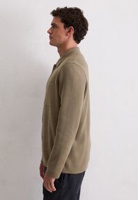 Lichtbruin, gestructureerde shirtjack met een kraag en een borstzak, met knoopsluiting en lange mouwen, gedragen over donkere broeken. Zijkant profielweergave.