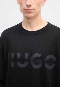 Juodas didelis medvilninis marškinėliai, su dideliu matinio juodo spalvos "HUGO" spaudu priekyje ir apvaliu kaklu.
