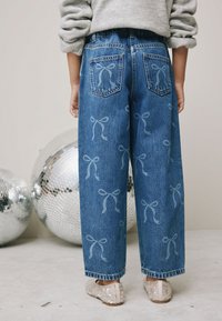 Blå denim jeans med vida ben har ljusblå rosettmönster, bakfickor och en resår i midjan. Matchas med paljettklädda ballerinaskor.