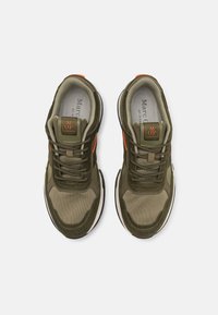 Vihreä oliivin- ja beige värinen kenkäpari, jossa on oranssit yksityiskohdat ja "M OP" -logo kielen päällä, kuvattuna ylhäältä valkoisella taustalla.