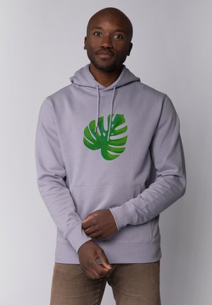 Man die een lichtpaarse hoodie draagt met een groene monstera-bladprint, staand met gevouwen handen tegen een effen lichtgrijze achtergrond.
