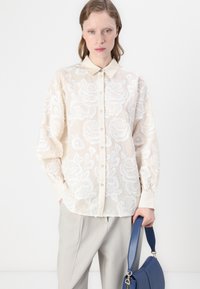 Blusa di crema trasparente con ricami floreali bianchi, chiusura a bottoni sul davanti, colletto a punta e maniche lunghe, abbinata a pantaloni grigi e una borsa blu.