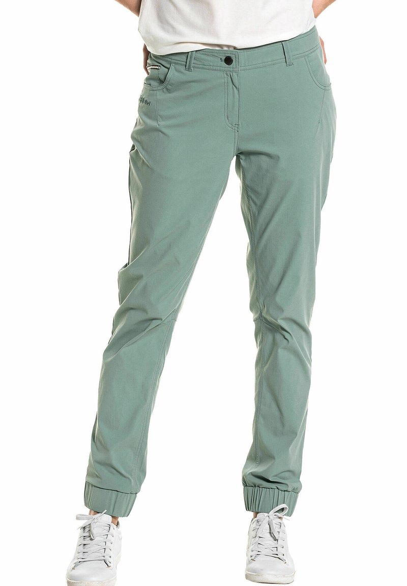 Personne portant un pantalon fuselé vert clair avec des poignets élastiques et des baskets blanches, debout les mains dans les poches.