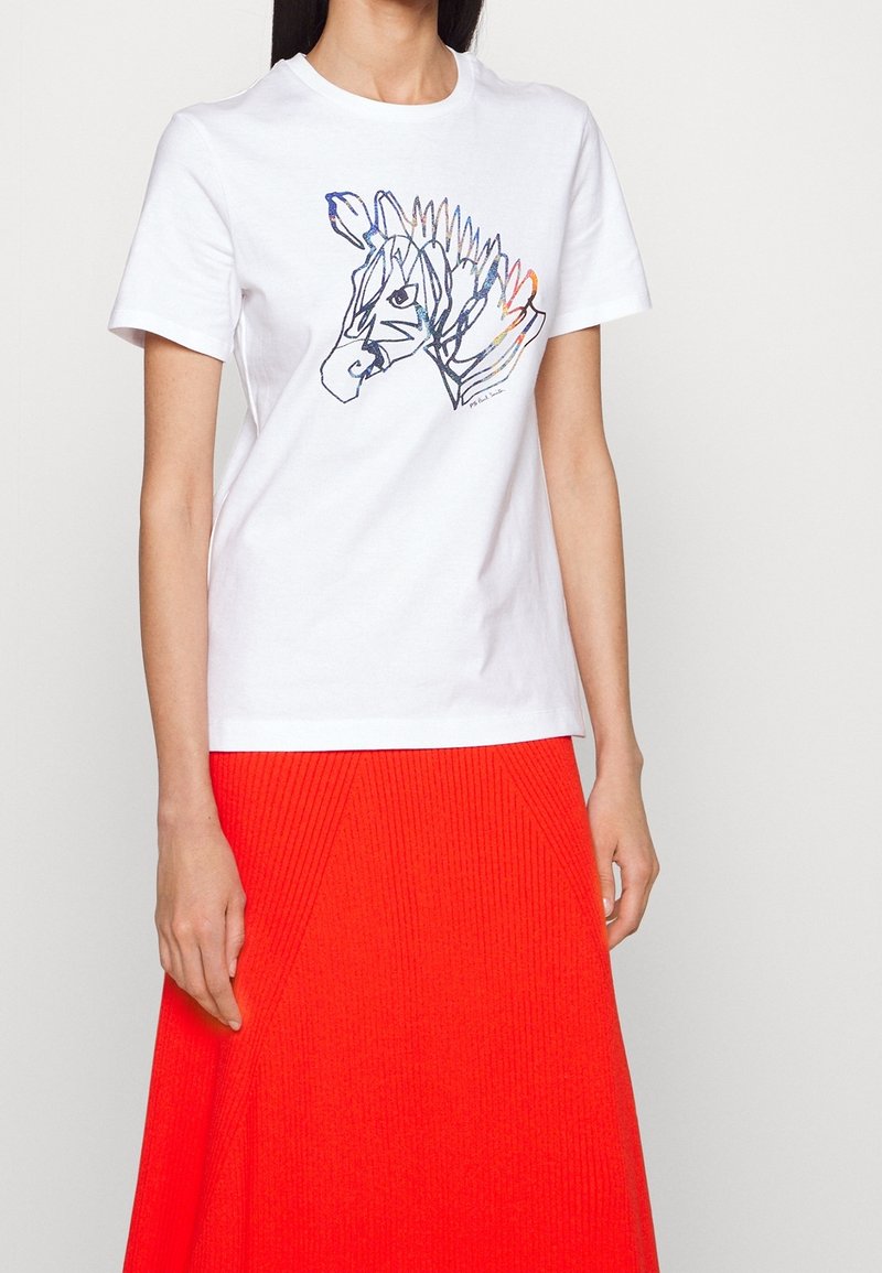 Kobieta ubrana w biały T-shirt z krótkim rękawem z kolorowym, abstrakcyjnym wzorem głowy zebry, w połączeniu z jasnoczerwoną, prążkowaną spódnicą.