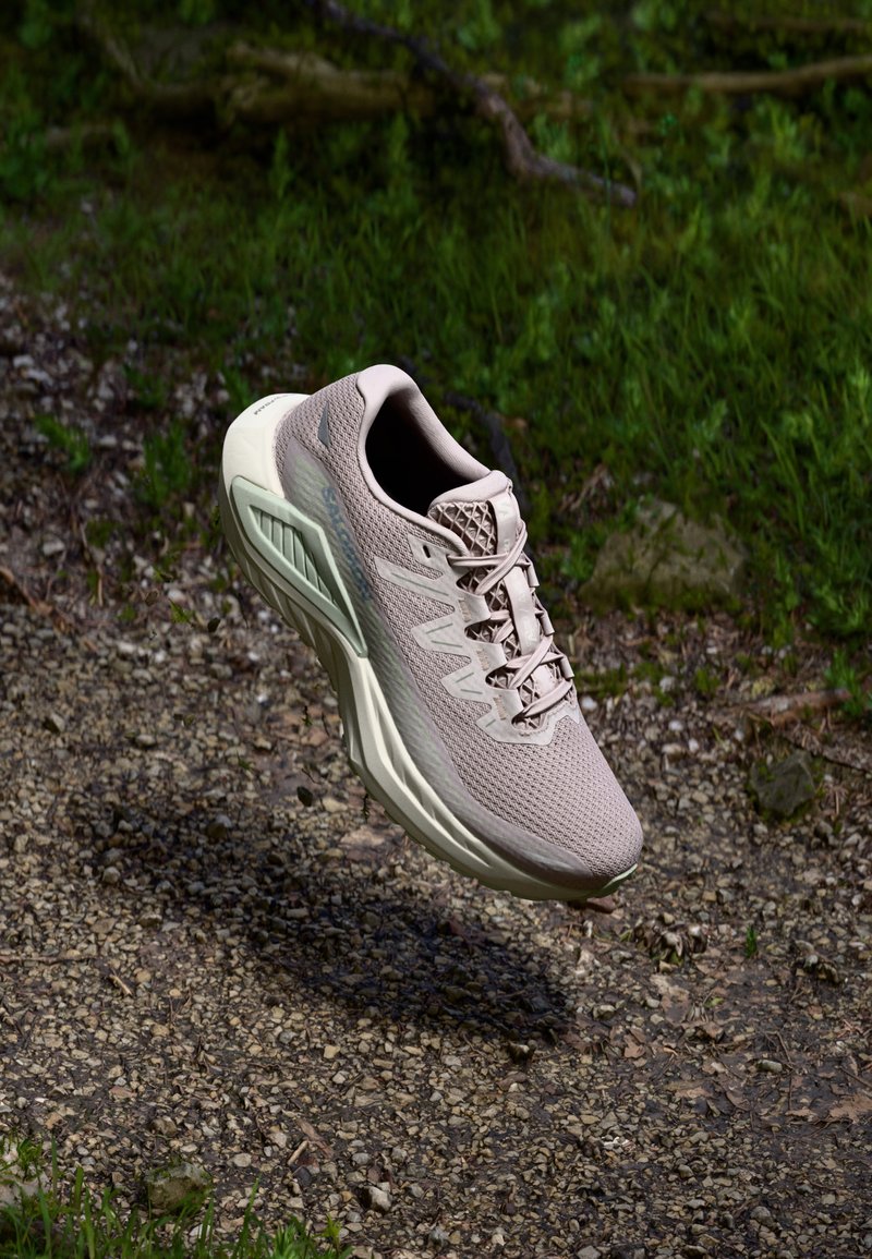 Chaussure de course beige clair avec une tige en maille respirante et un col rembourré. Semelle blanche épaisse avec des accents verts, mettant en valeur un design texturé.