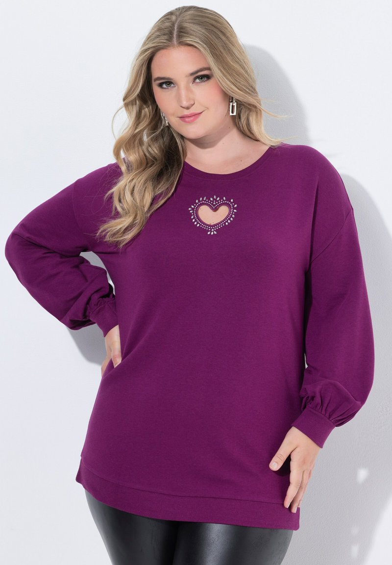 Donna con lunghi capelli biondi che indossa una felpa viola con un'apertura a forma di cuore decorata con strass, abbinata a pantaloni neri.