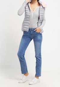 Gilet imbottito grigio leggero con cappuccio rosa, indossato sopra una maglietta grigia a maniche lunghe; abbinato a jeans blu e sneakers bianche.