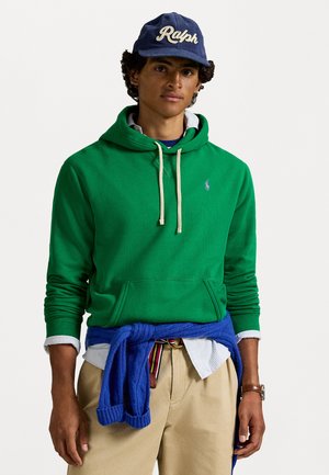 Groene katoenen hoodie met witte trekkoorden en een klein paars logo. Gelaagd over een blauwe sweater, gecombineerd met beige broeken en een blauwe pet.