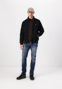 Svart zip-up jakke med ribbede mansjetter, en gul logo på brystet, brukt over en mørk skjorte, parret med blå jeans og svarte sko.