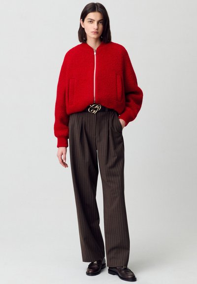 Giacca bomber rossa con texture, chiusura frontale con zip e due tasche, abbinata a pantaloni a vita alta marroni con strisce e scarpe scure.