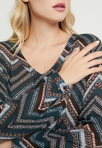 Haut à motifs avec un décolleté en V, combinant des rayures en zigzag bleu sarcelle, marron, rose et blanc, des poignets à boutons et une texture de tissu douce.