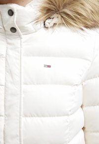 Vit puffjacka med huva prydd med päls, med horisontella quiltade sektioner och en liten logotyp i röd och blått på framsidan.