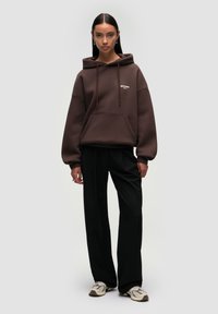 Brown hoodie with a front pocket, drawstring hood, and logo detail. Paired with black trousers and patterned sneakers.