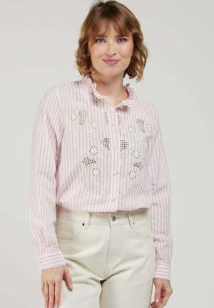 Femme aux cheveux courts et ondulés, portant un chemisier à rayures roses et blanches avec des découpes florales et un pantalon blanc, debout devant un fond uni.