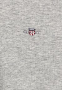 Pull en coton gris avec un tricot texturé, arborant un petit logo brodé en rouge, blanc et bleu sur la poitrine gauche.