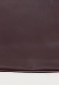 Σκούρο μπορντό δερμάτινο πουγκί με λεία υφή, διαθέτοντας ανάγλυφο λογότυπο και μινιμαλιστικό σχεδιασμό. Χωρίς ορατό υλικό ή διακοσμητικά στοιχεία.