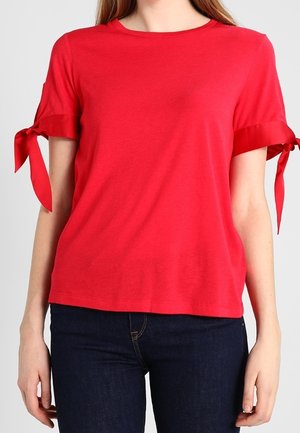 Femme portant un haut rouge à manches courtes avec des nœuds noués sur les manches et un jean bleu foncé, debout devant un fond uni.