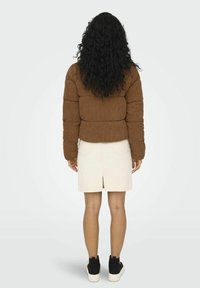 Bruine corduroy pufferjack, strakke taille, gedragen over een licht crèmekleurige rok. Zwarte sneakers met witte zolen. Lange krullende zwarte haren zichtbaar.