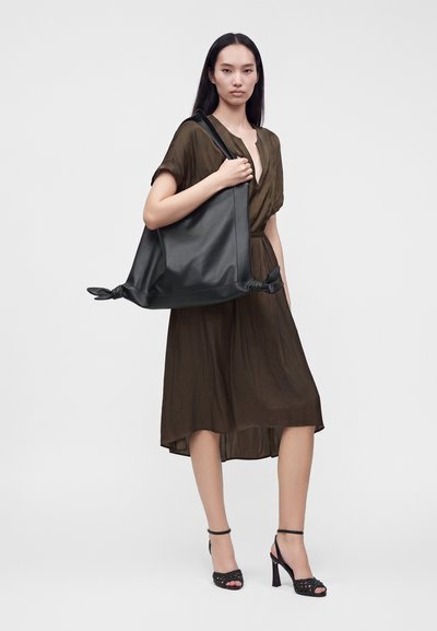 Femme portant une robe marron et des sandales noires à talons hauts, tenant un grand sac cabas noir avec des coins noués sur une épaule.