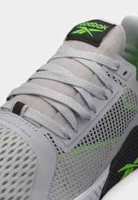 Κοντινό πλάνο ενός γκρι και πράσινου αθλητικού παπουτσιού Reebok που δείχνει το υφασμάτινο πλέγμα με υφή, τα κορδόνια και το λογότυπο στη γλώσσα και στην πλευρική σόλα.