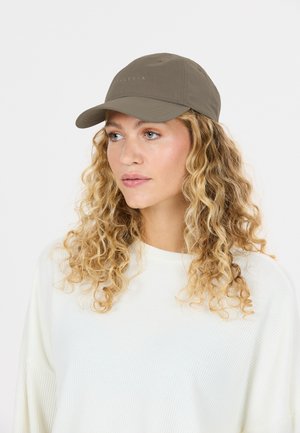 Femme blonde aux cheveux bouclés porte une casquette de baseball taupe et un pull blanc ample, regardant légèrement à droite sur un fond blanc uni.