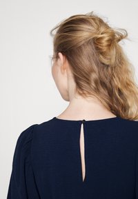 Top blu navy con apertura a chiave sul retro e chiusura a bottone, texture morbida e dettaglio delle spalle arricciato. I tessuti sembrano leggeri.