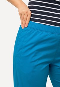 Turkosa byxor med elastisk midja, med en slät yta och sidofickor, delvis täckta av en hand på höften. Randig topp synlig ovanför.