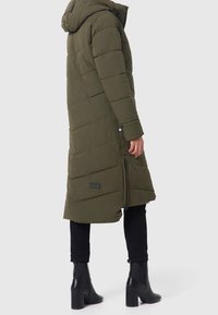 Lång olivgrön puffjacka med huva, med en kviltad design och sidogympar, kombinerad med svarta åtsittande byxor och ankelboots.