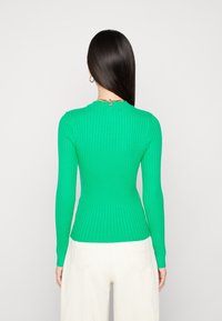 Top verde a maniche lunghe a coste con design aderente e scollo rotondo, caratterizzato da tessuto testurizzato e un dettaglio sottile sulla schiena. Pantaloni bianchi.
