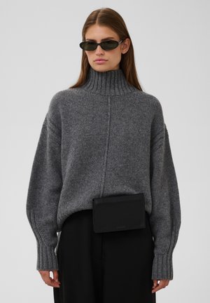Femme portant des lunettes de soleil foncées, un épais pull à col roulé gris, un pantalon noir et une banane noire, debout devant un fond uni.