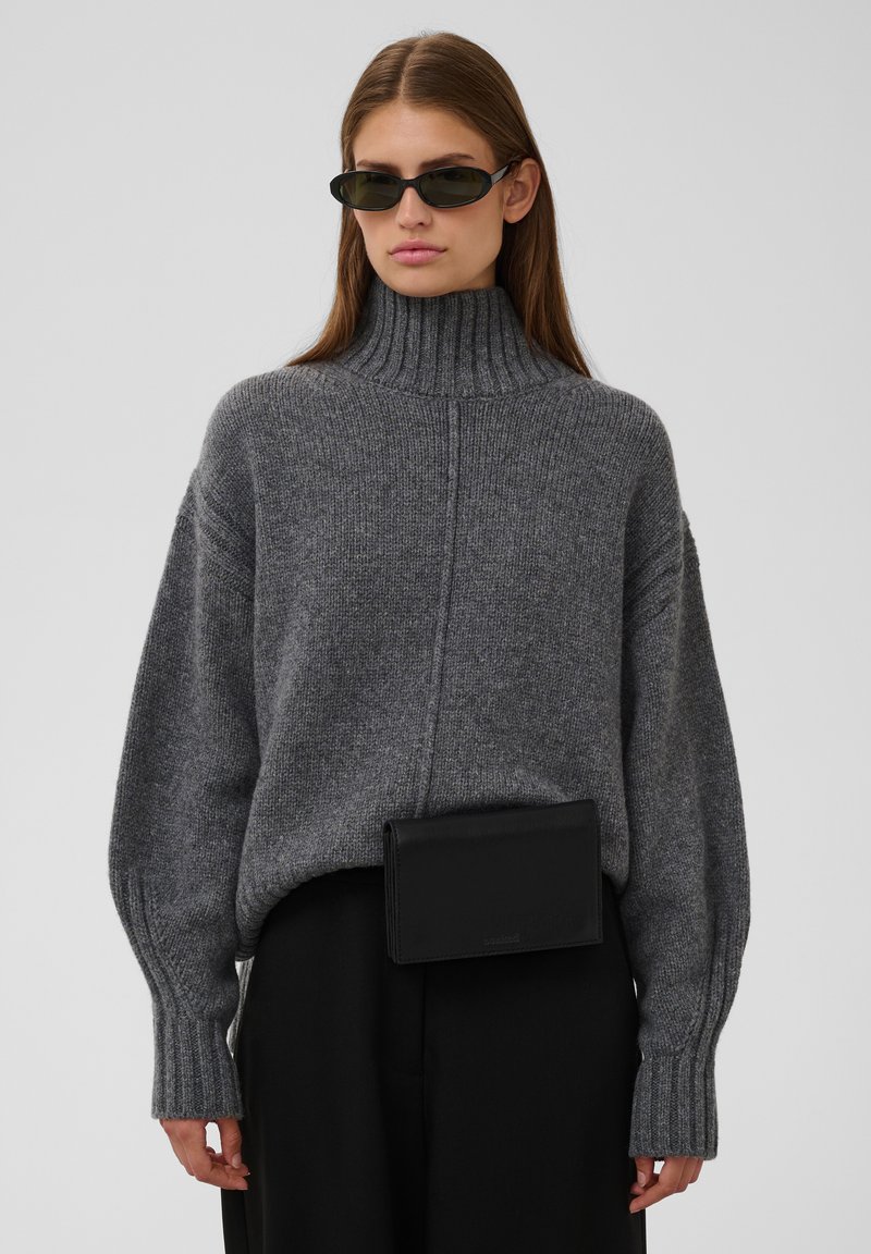 Femme portant des lunettes de soleil foncées, un épais pull à col roulé gris, un pantalon noir et une banane noire, debout devant un fond uni.