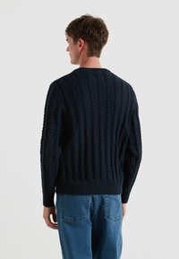 Navyblauwe gebreide trui met een gestructureerd kabelpatroon, voorzien van geribbelde manchetten en zoom, van achteren weergegeven met een relaxed-fit spijkerbroek.